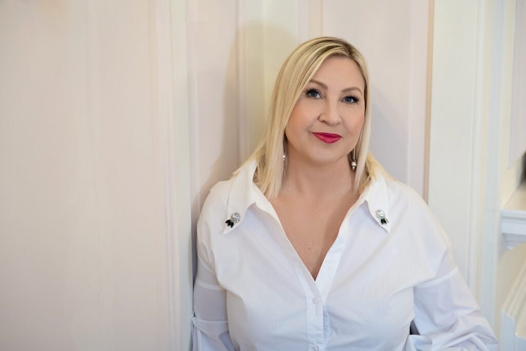 Tracy Fensome Headshot with Tracy leaning against a wall facing toward camera, blonde hair, red lipstick, white open neck shirt