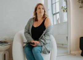 Image of Stephanie Barnard, Your Beauty Business Bestie, wearing navy blue vest, with pale blue cardigan and jeans sitting on white low back armchair with arch way to right behind her