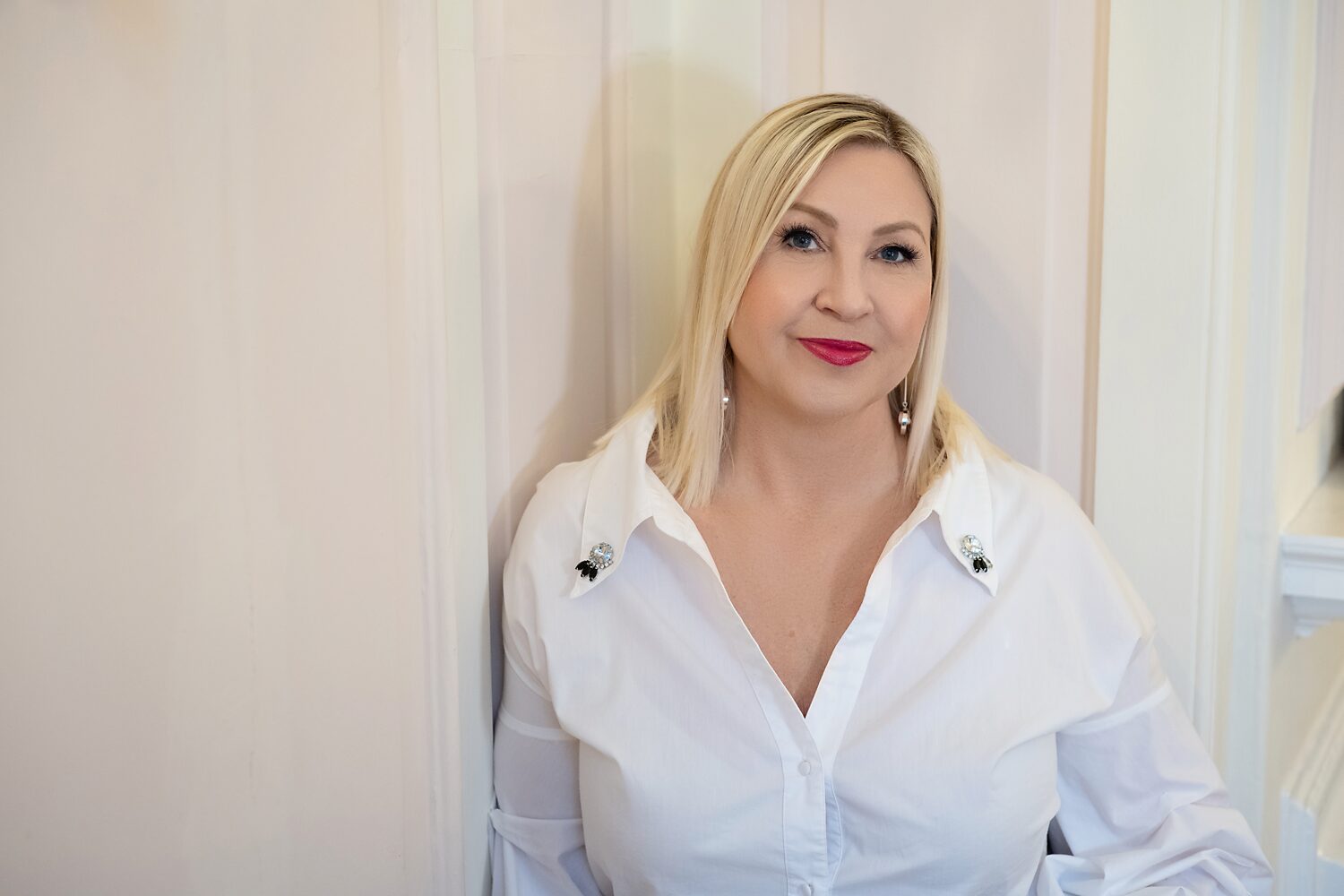 Tracy Fensome Headshot with Tracy leaning against a wall facing toward camera, blonde hair, red lipstick, white open neck shirt