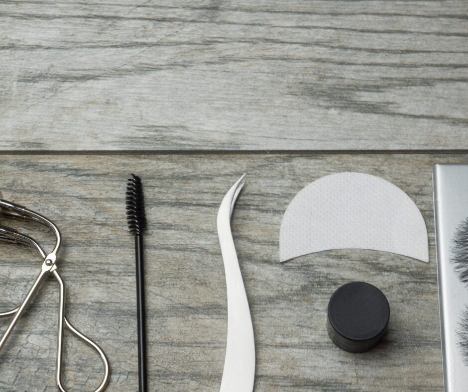 Lash Extension tools including tweezers, spoolie, under eye protection pads, and lash curlers laid on a natural wood background