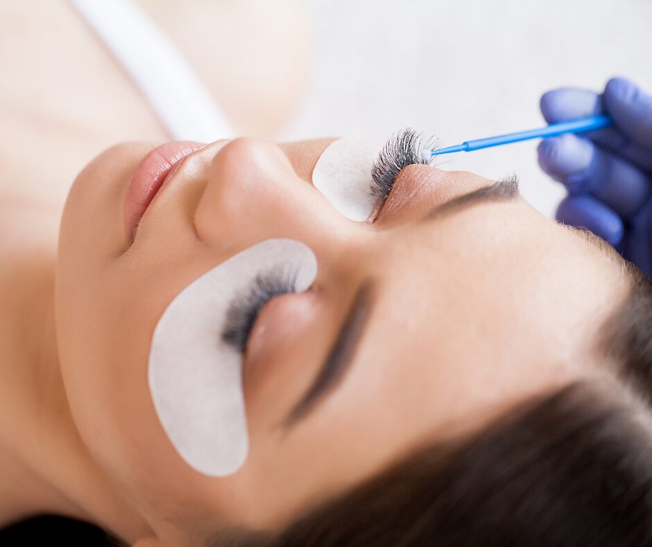 Lash extensions being applied to a client who lays on beauty bed with lash technician applying lash products to clients lash line. Client has dark hair, dark brows and wears under eye protection pads. For salonindustrynetworking.co.uk